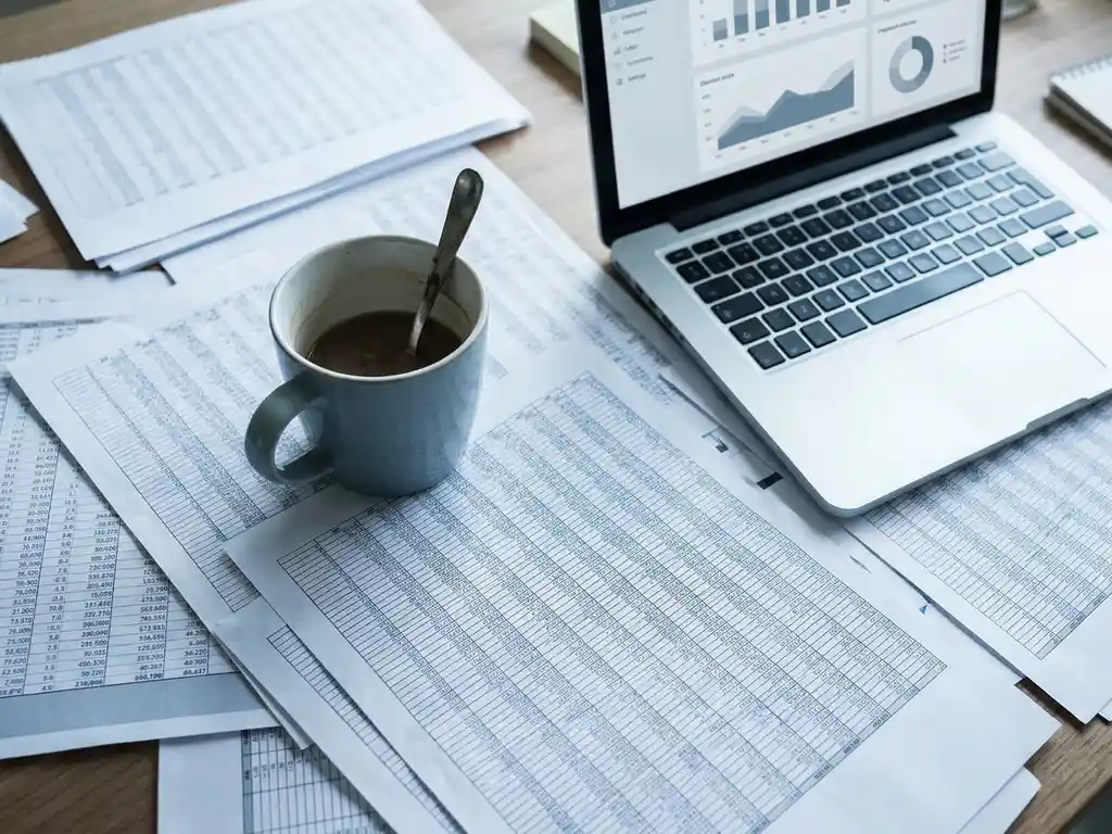 Cluttered desk flat lay with printed spreadsheet pages, cold coffee mug, and a laptop showing a clean dashboard in the corner.