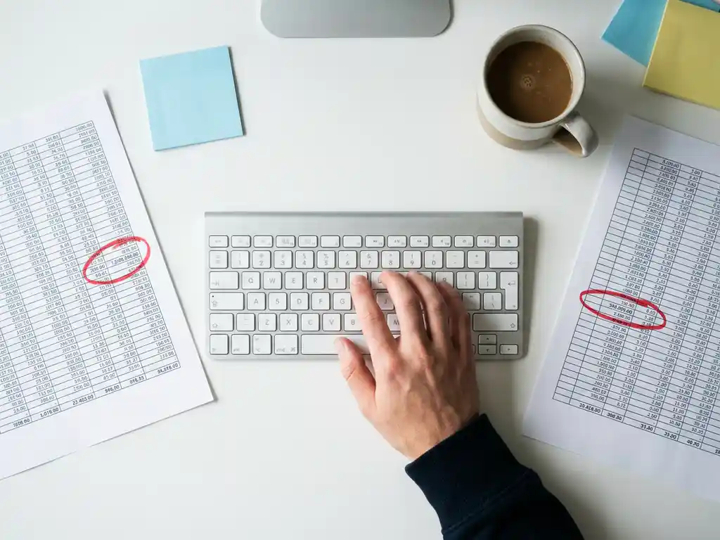 Trader's hand over keyboard beside spreadsheet printouts with red-circled mismatched figures, sticky notes, and cold coffee on a white desk.